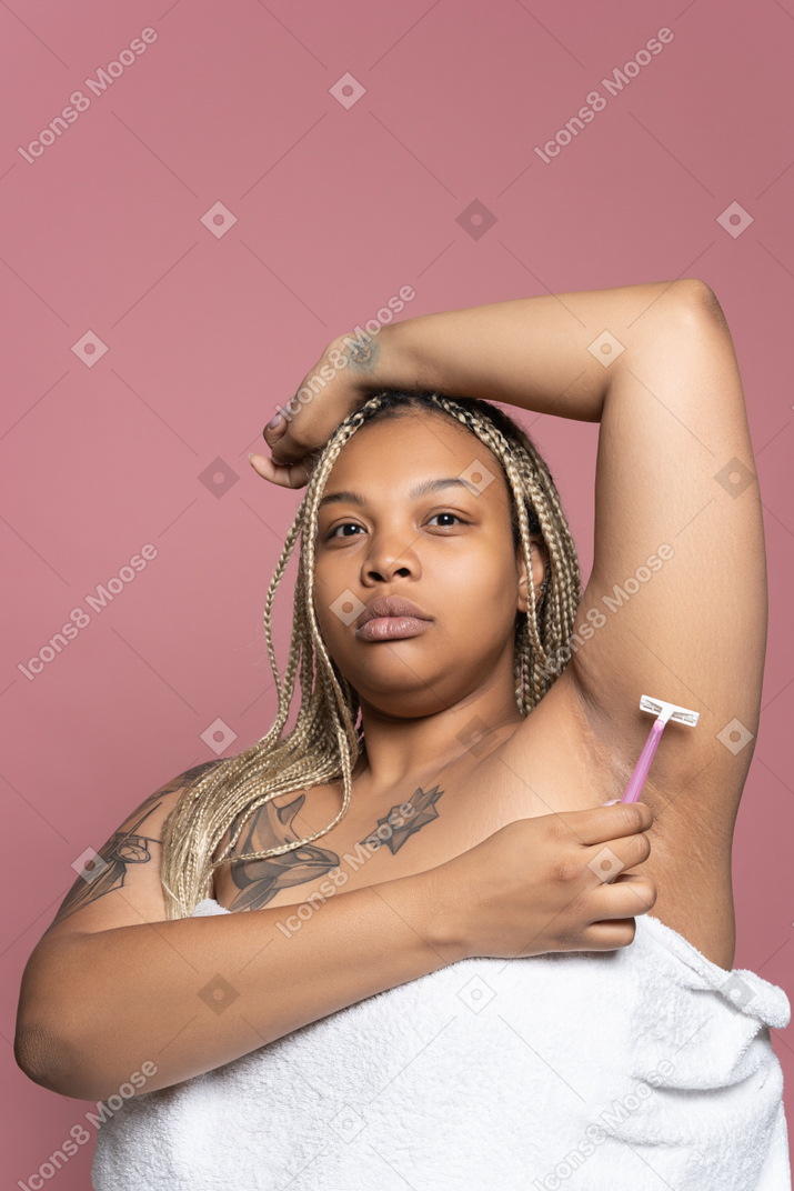 Plum afro woman wrapped in white towel shaving underarm