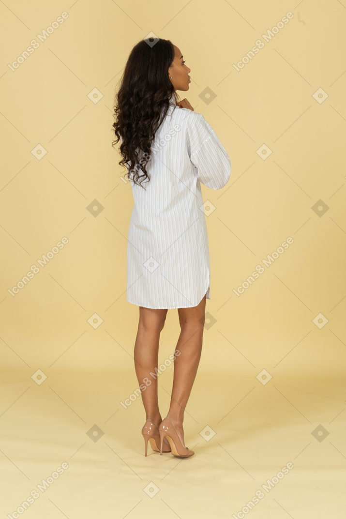 Three-quarter back view of a dark-skinned young female in white dress adjusting her collar