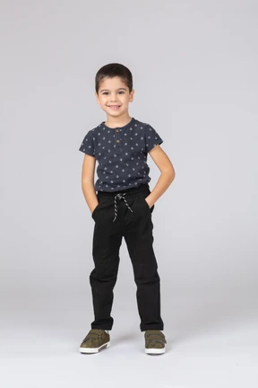 Front view of a happy boy in casual clothes posing with hands in pockets and looking at camera