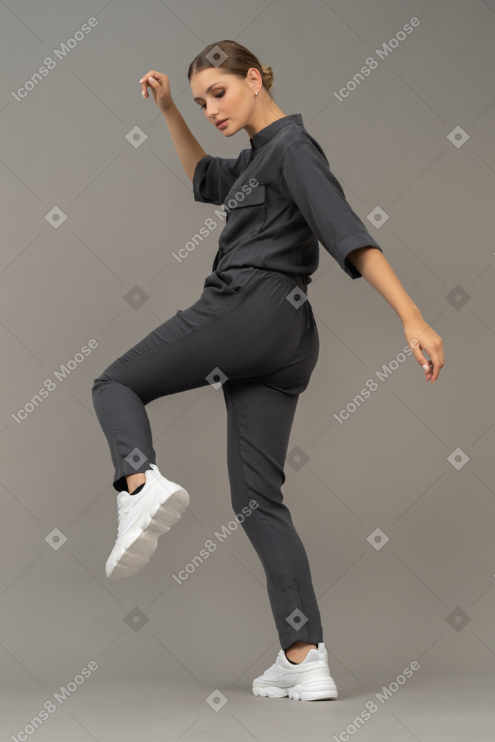 Side view of a young woman in a jumpsuit outstretching her arm & raising leg