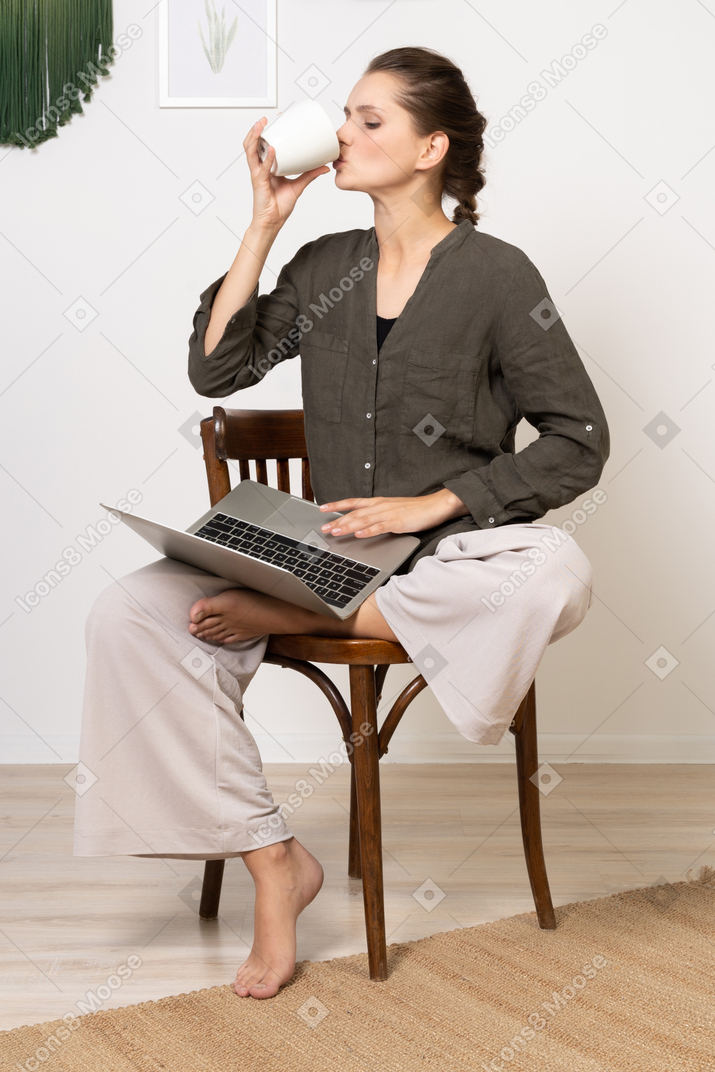 Vista frontal de uma jovem vestindo roupas de casa, sentada em uma cadeira com um laptop e bebendo café