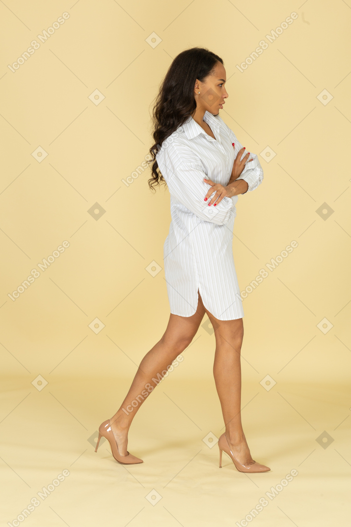 Вид сбоку идущей темнокожей молодой девушки в белом платье, скрещивающей руки