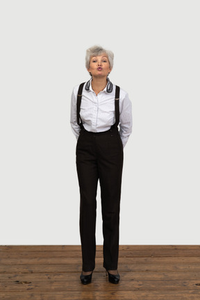 Front view of an old funny female in office clothes grimacing with her hands behind back sending a kiss