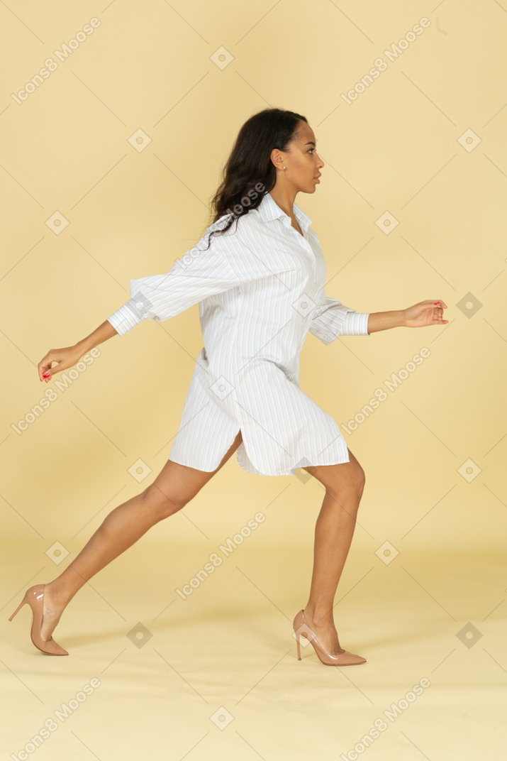 Vista lateral de uma jovem mulher de pele escura em um vestido branco dando um grande passo