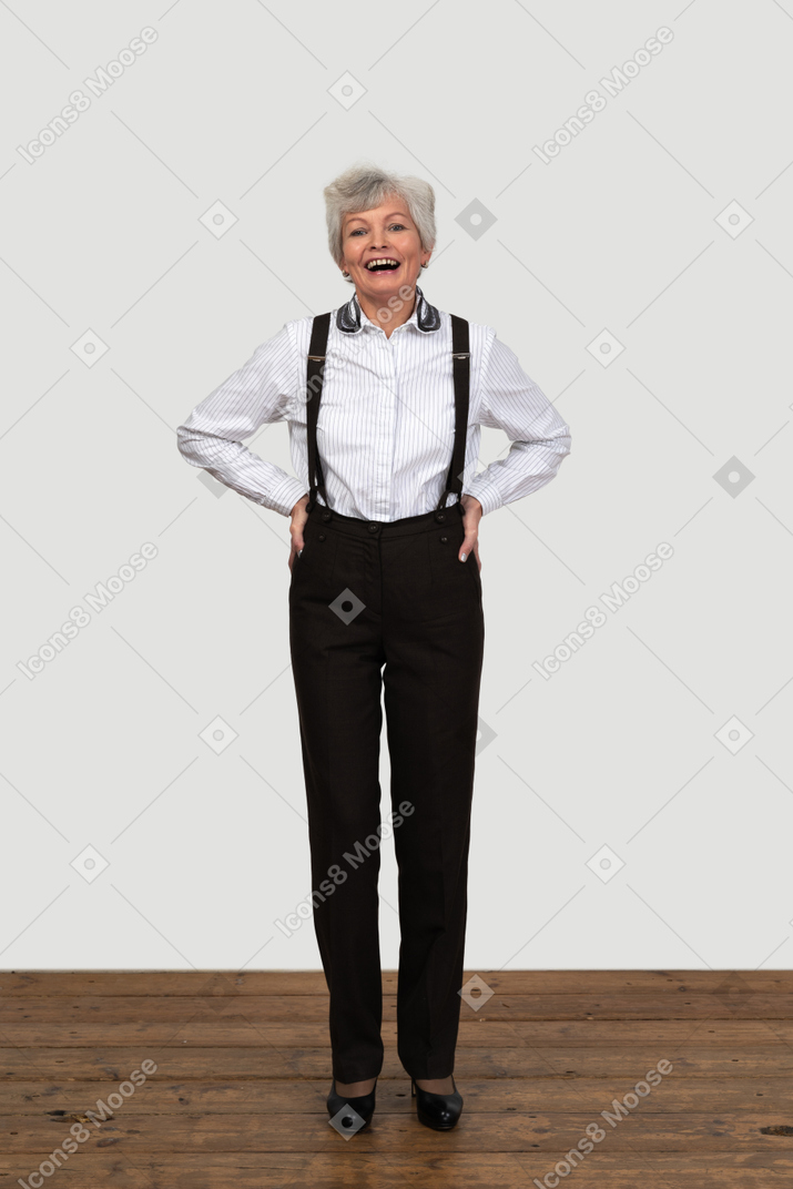 一个老妇人站在木地板上穿着带吊带裤的裤子，笑着把手放在臀部上