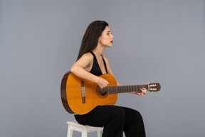 Three-quarter view of a sitting young lady in black suit holding the guitar and looking aside