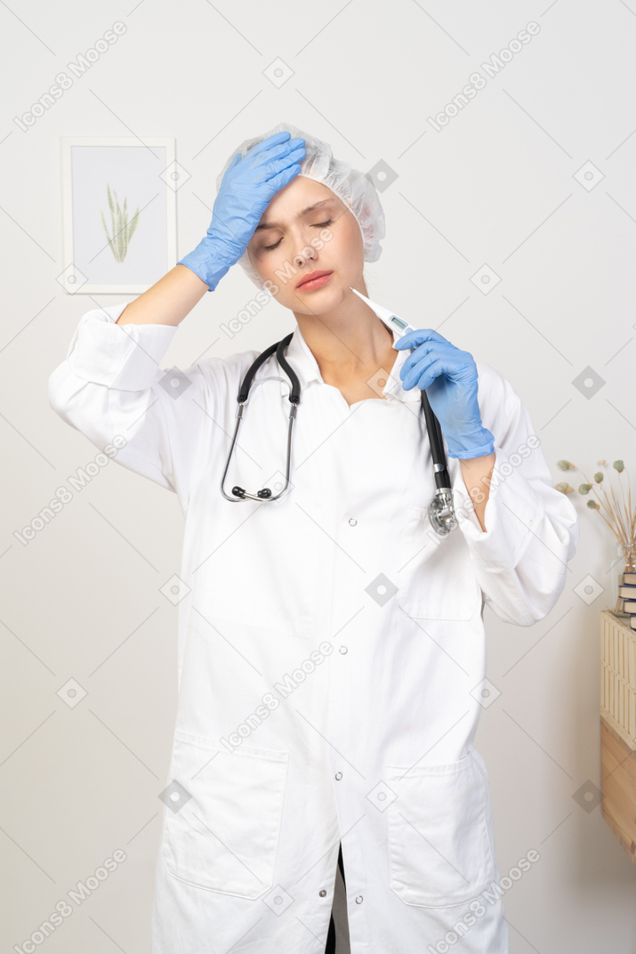 Vista frontal de una joven doctora con estetoscopio sosteniendo el termómetro y tocando la cabeza