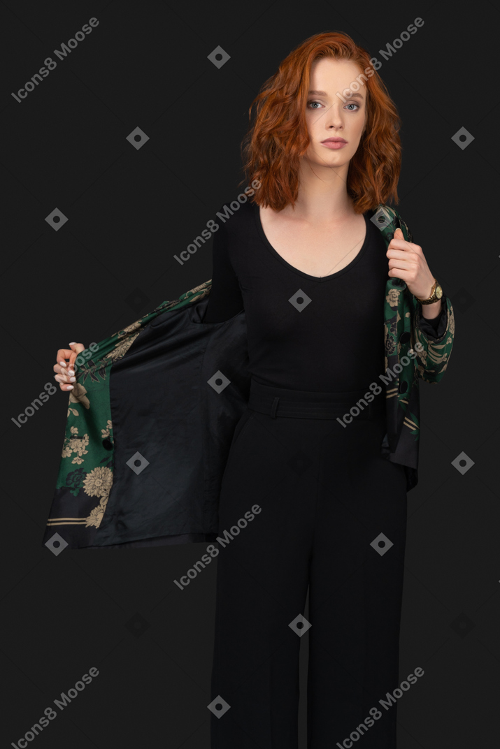 Young woman in black top and pants looking into camera and taking her jacket off