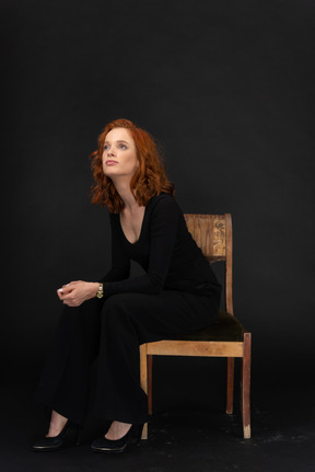 Молодая женщина в черном наряде сидит на стуле и смотрит вверх