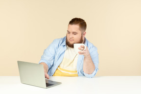 Молодой человек с избыточным весом работает на ноутбуке и пьет чай