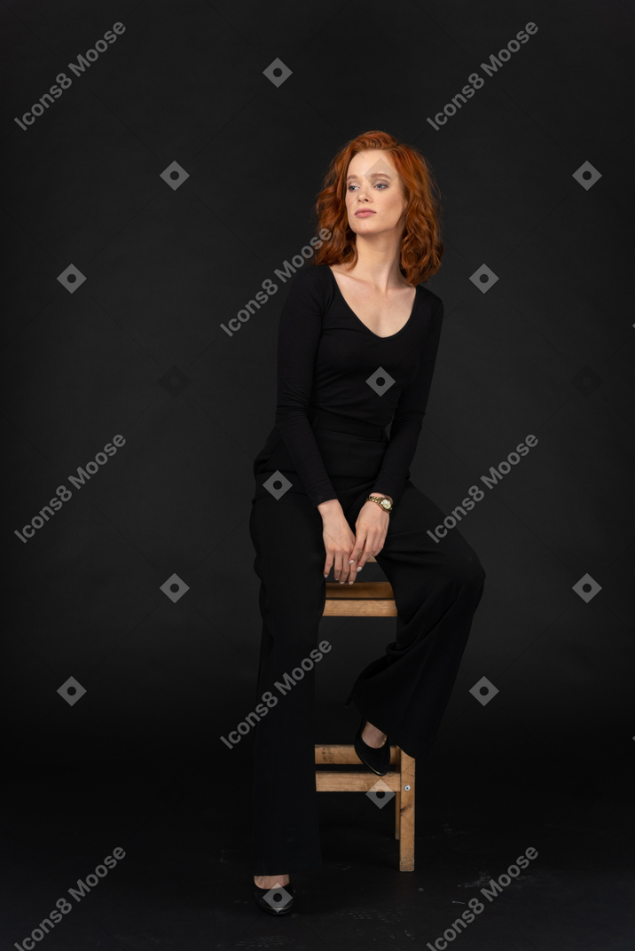 A frontal view of the cute woman dressed in black and sitting on the chair on the dark background