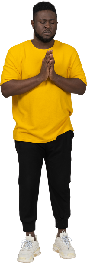 Vista frontal de um jovem de pele escura orando em uma camiseta amarela de mãos dadas