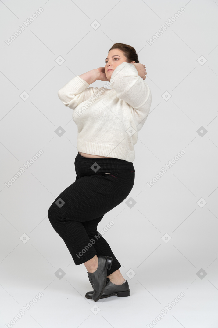 Mujer regordeta en suéter blanco de pie con las manos detrás de la cabeza
