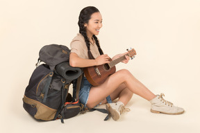 Sonriente joven asiática sentada cerca de la mochila y tocando la guitarra