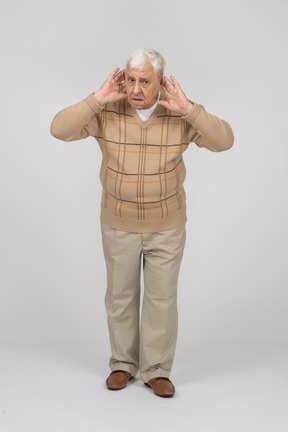 Front view of an old man in casual clothes holds hands close to ears
