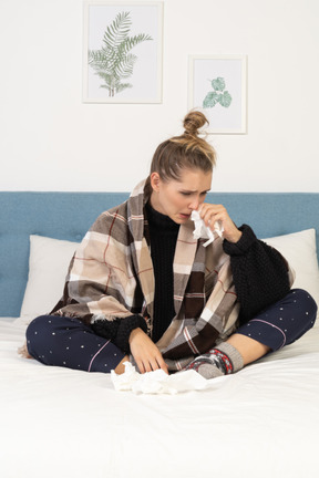 Front view of an ill young lady in pajamas wrapped in checked blanket in bed blowing her nose