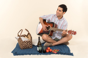 Junger kaukasischer kerl, der nahe picknickkorb auf der decke sitzt und gitarre spielt