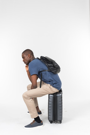 Pensive male tourist sitting on grey luggage