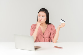 Duvidando jovem mulher asiática segurando o cartão do banco ao fazer compras on-line