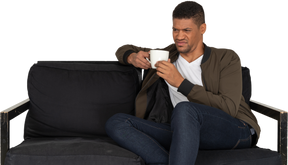 Front view of a young grimacing man sitting on a sofa with a cup of coffee