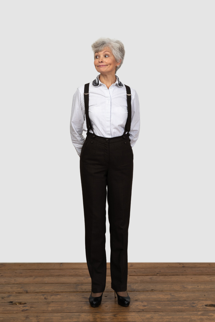 Front view of an old smiling female in office clothes grimacing with her hands behind back looking aside