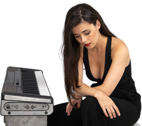 Corpo inteiro de uma jovem triste vestida de preto sentada ao piano e segurando as pernas de mãos dadas