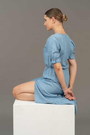 Back-side view of young woman in blue dress sitting on cube