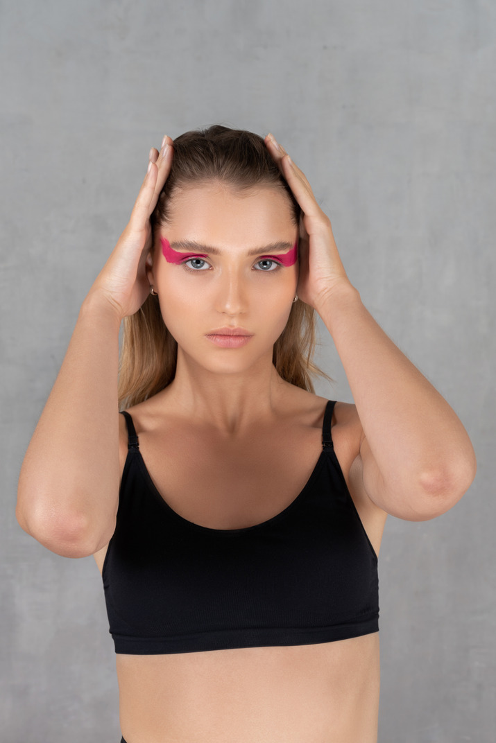 Front view of a young woman with bright pink eye make-up looking at camera & touching head
