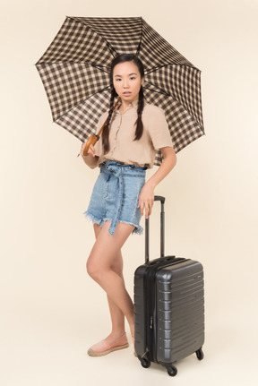 Mujer joven seria con paraguas y maleta mirando a la cámara