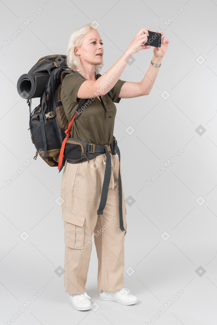 Mature female tourist carrying heavy backpack and making photos with smartphone
