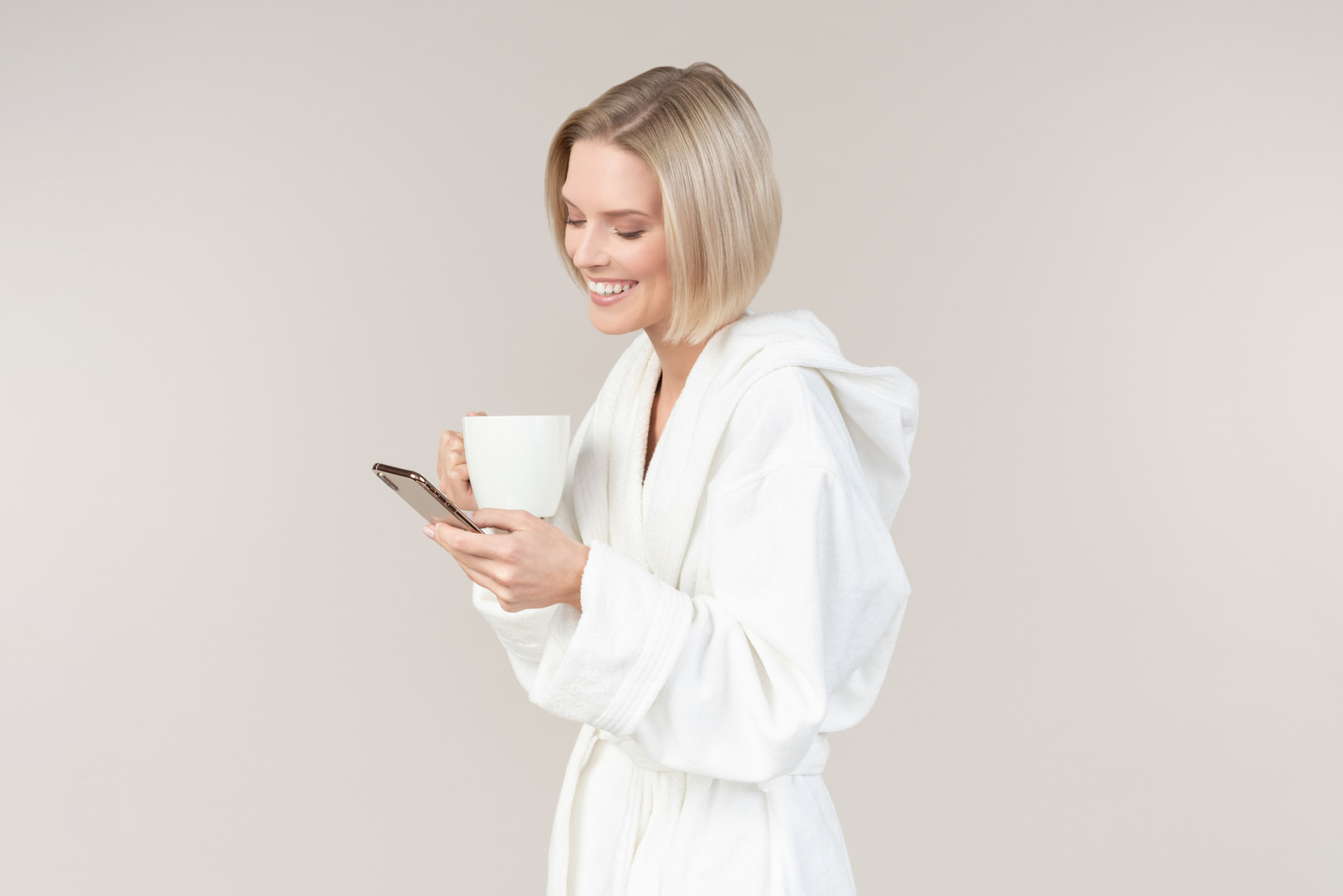 Smiling young woman in bathrobe using phone and having coffee