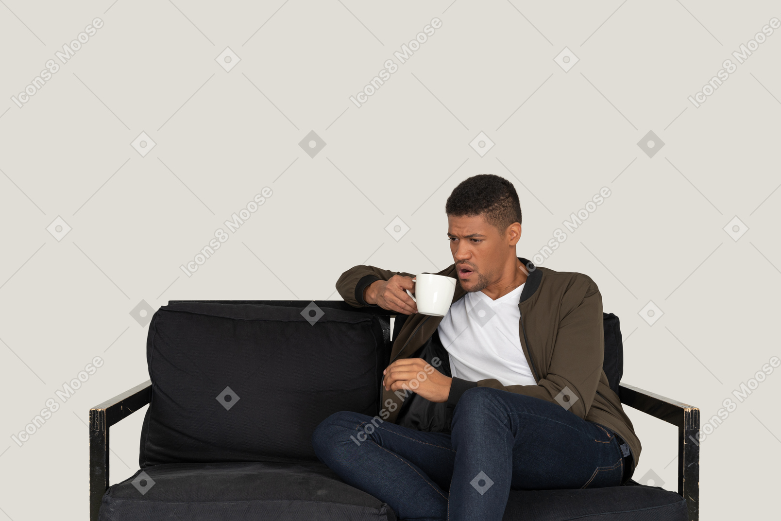 Front view of a young displeased man sitting on a sofa with a cup of coffee