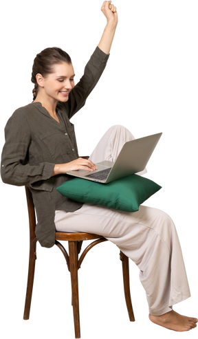 ノートパソコンと手を上げて椅子に座って家庭服を着ている若い女性の4分の3のビュー