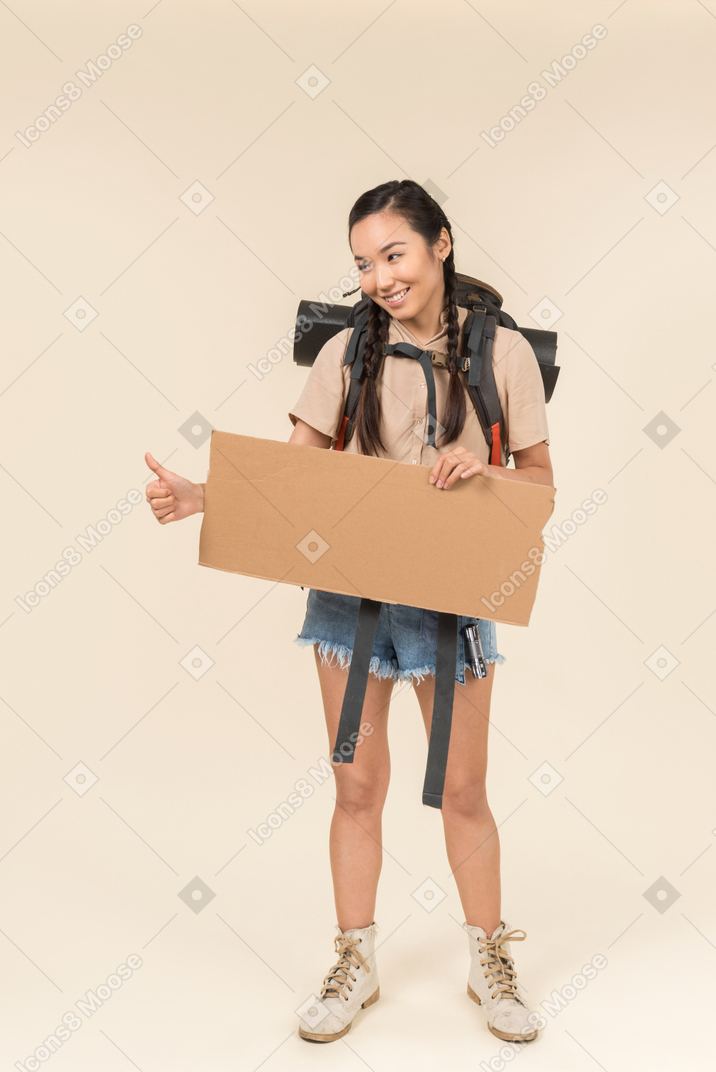 Junge weibliche anhalterin, die papierkarte hält und sich daumen zeigt