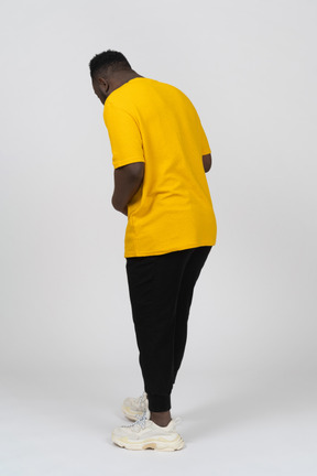 Three-quarter back view of a young dark-skinned man in yellow t-shirt touching stomach & looking down