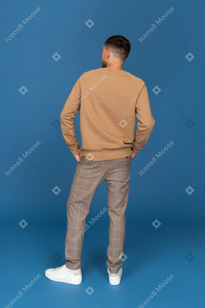 Молодой человек с руками в карманах стоит спиной к камере