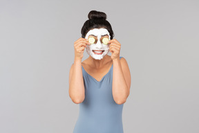 Woman with white facial mask applying cucumber slices on eyes