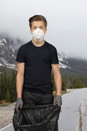 Man standing on a road in face mask and holding black trash bag