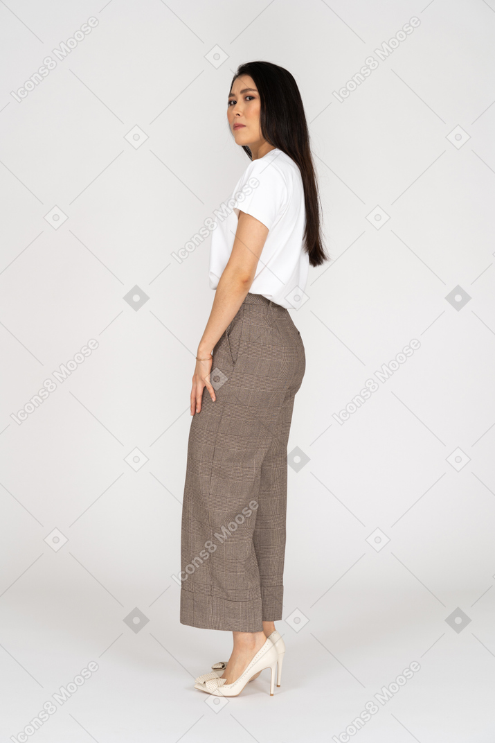 Vista laterale di una giovane donna sospettosa in calzoni e t-shirt che guarda l'obbiettivo
