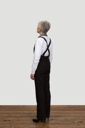Vista de três quartos de uma mulher idosa com suspensórios, parada em um quarto vazio