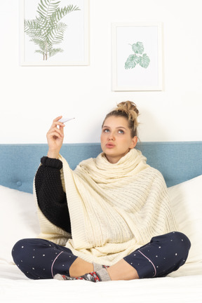 一位裹着白色毯子的年轻女士坐在床上，拿着温度计的前视图