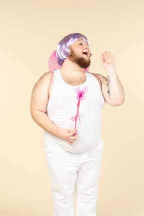 Young overweight man dressed as a fairy holding fairy wand and looking aside