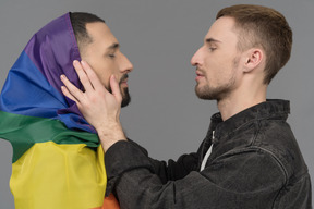 Nahaufnahme eines jungen mannes, der das gesicht eines anderen mannes hält, der in eine lgbt-flagge gehüllt ist