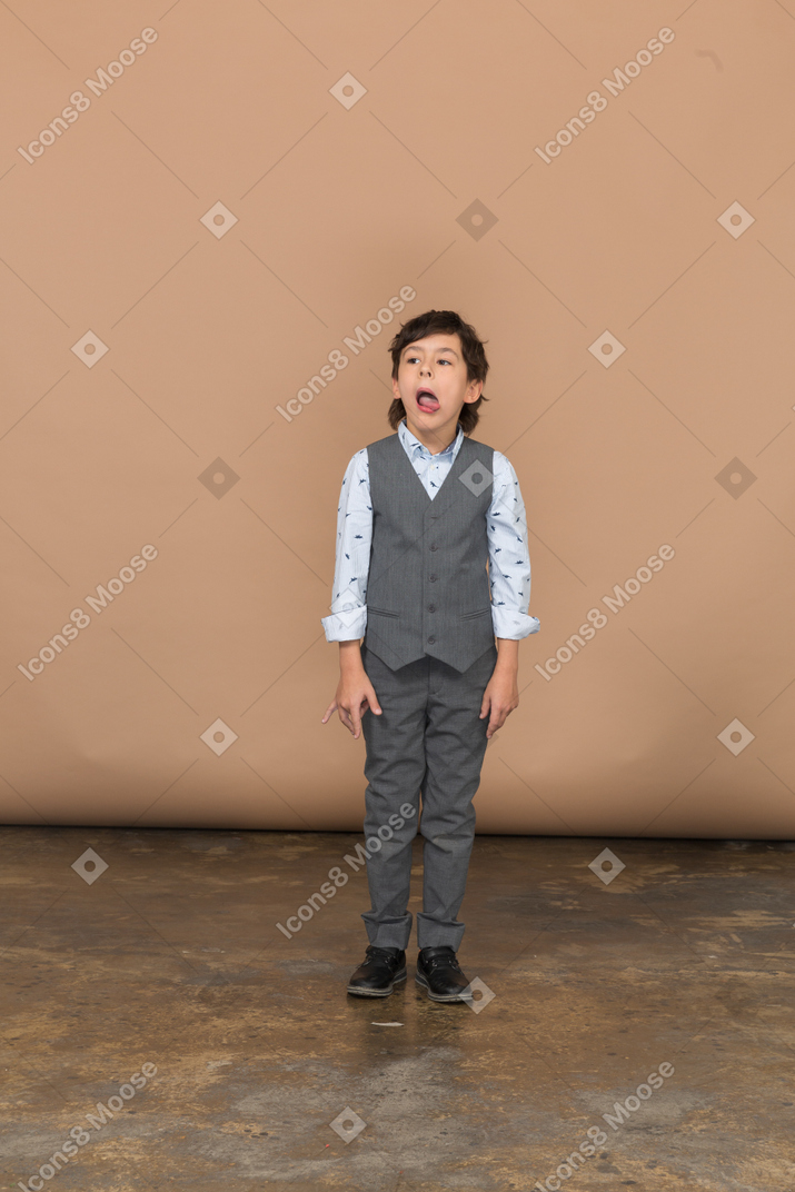 Vista frontale di un ragazzo carino in abito grigio che fa smorfie e mostra la lingua