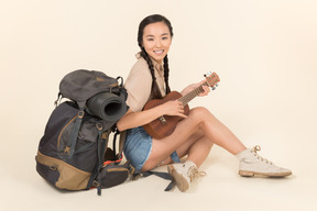 Smiling young asian girl sitting near backpack and playing the guitar