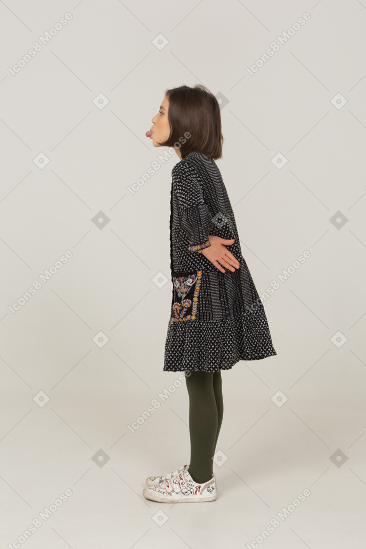 Side view of a funny little girl in dress showing tongue and putting hands on hips