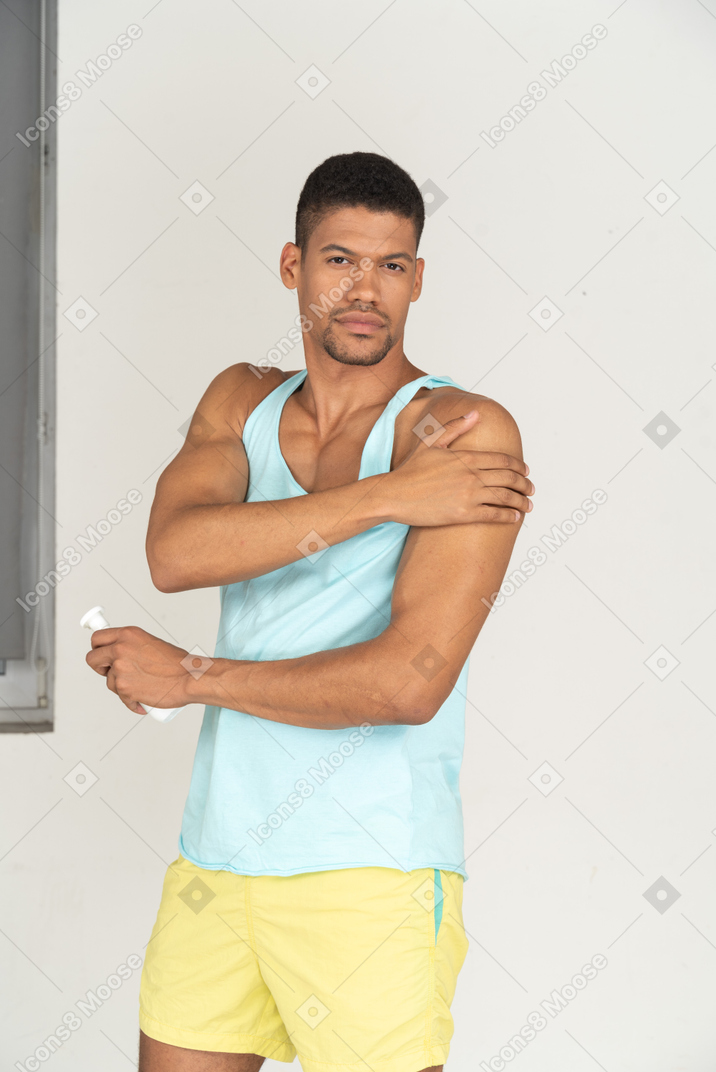 A man in a blue tank top and yellow shorts putting on a sunscreen