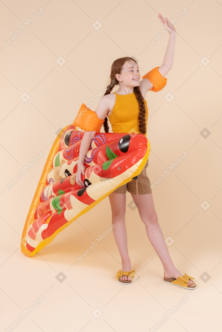 Teenage girl wearing arm floats, holding pizza mattress and waving