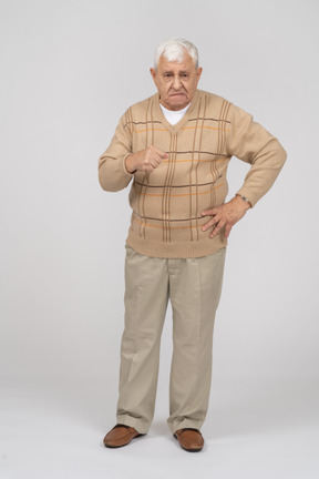 Front view of an old man in casual clothes standing with hand on hip and explaining something