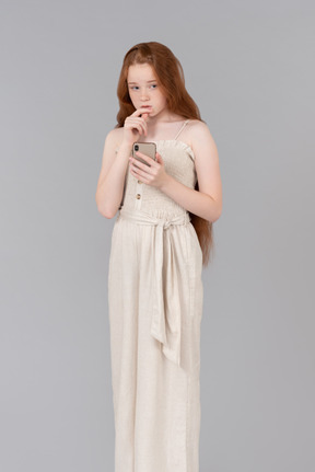 Pensive teenage girl in beige overalls using phone